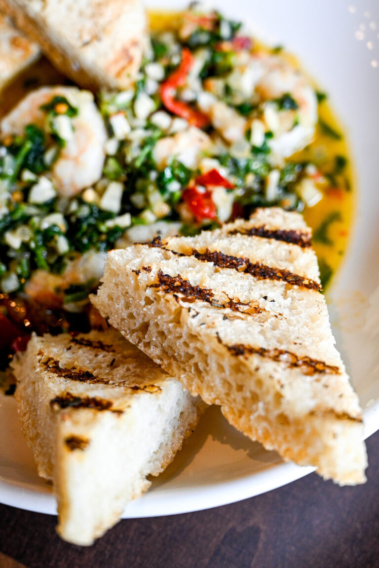 A close-up of a dish featuring three pieces of grilled bread arranged atop a vibrant mix of chopped herbs, diced vegetables, and shrimp in a light, flavorful broth. The bread shows distinct grill marks, adding a touch of rustic presentation to the meal.