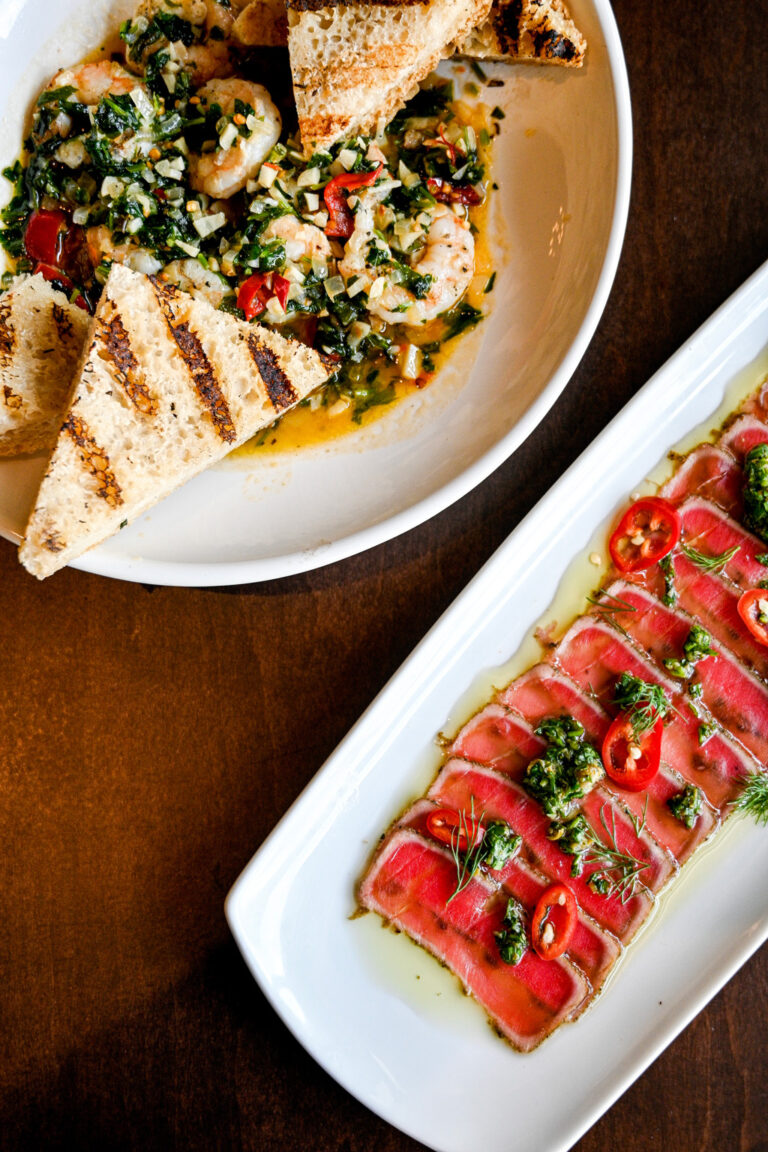 A plate of grilled shrimp with herbs, chopped vegetables, and wedges of grilled bread next to another plate of thinly sliced seared tuna drizzled with oil and garnished with red chili slices and herbs. Both dishes are presented on a wooden table.