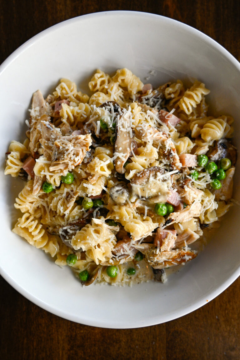 A white bowl filled with pasta, topped with pieces of chicken, peas, mushrooms, and a generous sprinkle of grated cheese. The pasta is arranged in a spiral shape and coated with a creamy sauce, set against a dark wooden background.