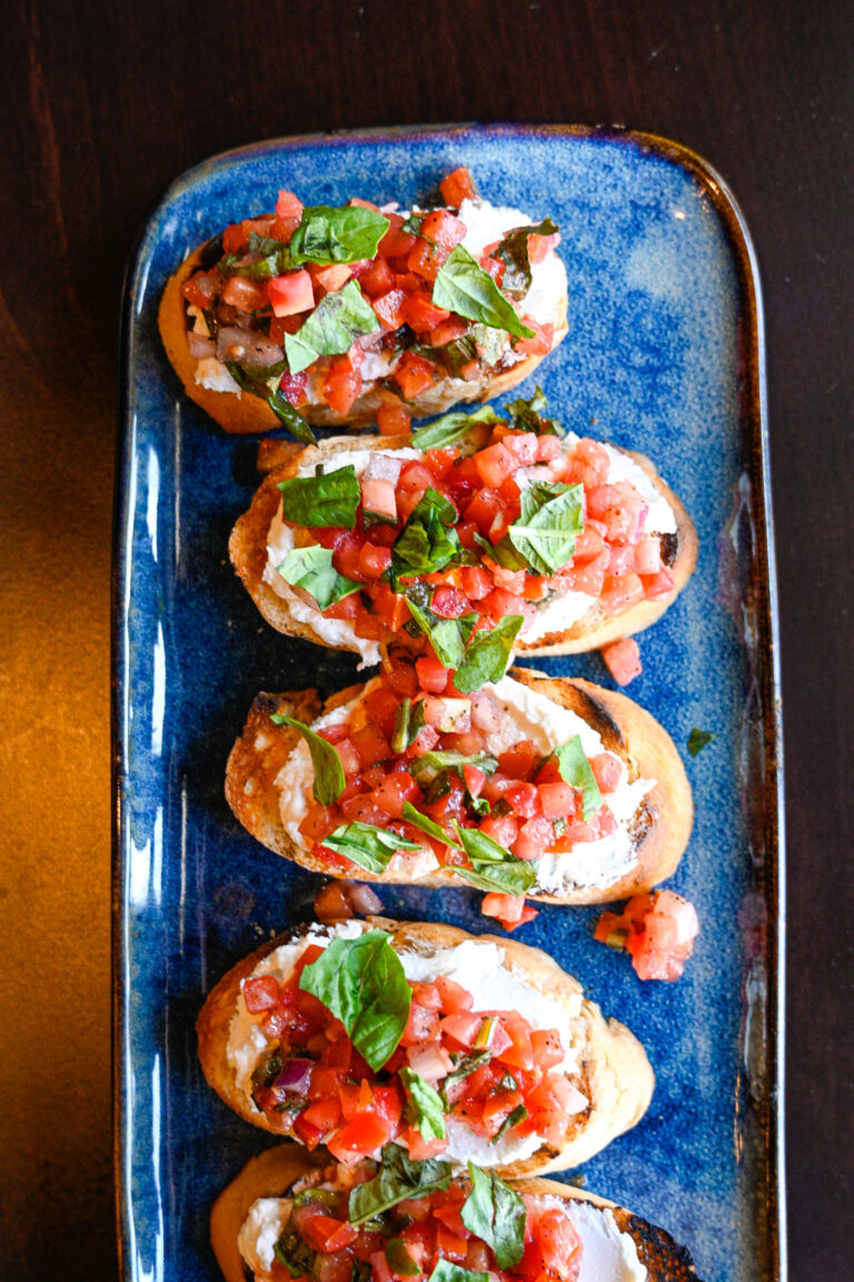 A rectangular blue platter holds several pieces of bruschetta. Each slice of toasted bread is topped with a layer of creamy cheese, diced tomatoes, and fresh basil leaves. The vibrant colors of the toppings contrast with the dark background surface.