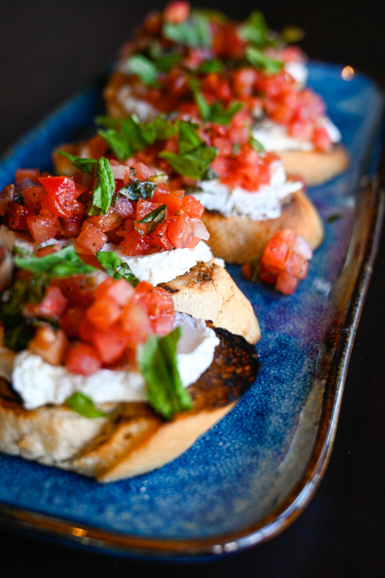 A close-up of four bruschetta pieces on a blue rectangular plate. The bruschetta features a toasted bread base topped with a generous layer of diced tomatoes, fresh basil, and a dollop of creamy ricotta cheese.