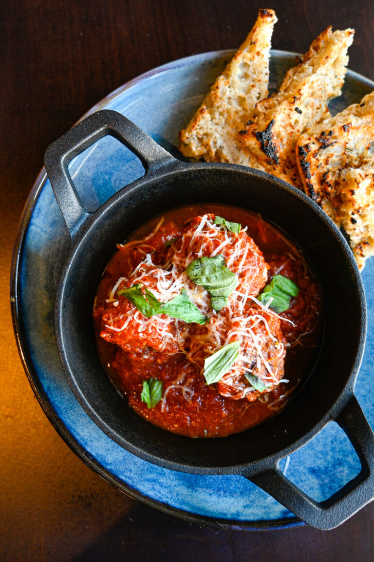 A black bowl filled with meatballs in a rich tomato sauce, topped with grated cheese and fresh basil, placed on a blue plate. Slices of toasted bread are arranged on the side of the plate, resting on a dark wooden surface.