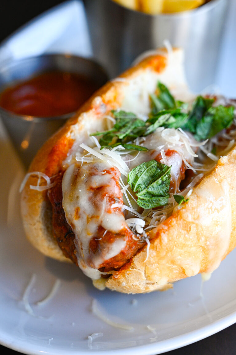 A close-up of a meatball sub on a white plate. The sub is topped with melted cheese, marinara sauce, and fresh basil leaves inside a crusty bun. A stainless steel container holding fries and a cup of ketchup is visible in the background.