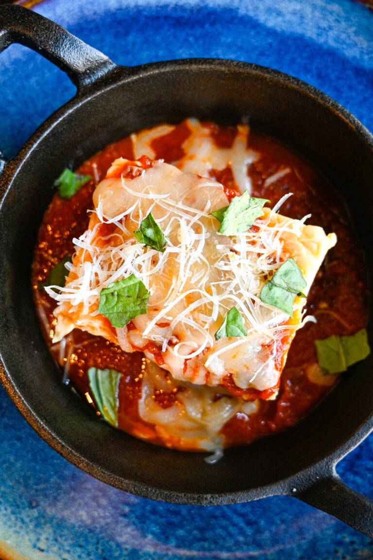 A close-up photo of a freshly baked lasagna in a black cast iron skillet, topped with melted cheese, grated Parmesan, and fresh basil leaves. The skillet is placed on a blue plate.