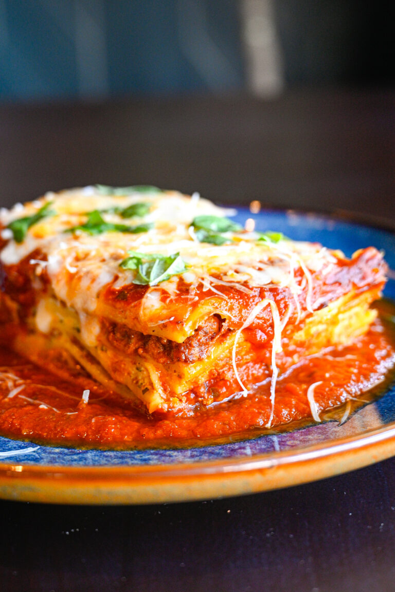 A close-up of a piece of lasagna on a plate. The lasagna has layers of pasta, meat, rich tomato sauce, and melted cheese, garnished with fresh basil leaves. Some sauce spreads around the base of the lasagna, adding a vibrant splash of red.