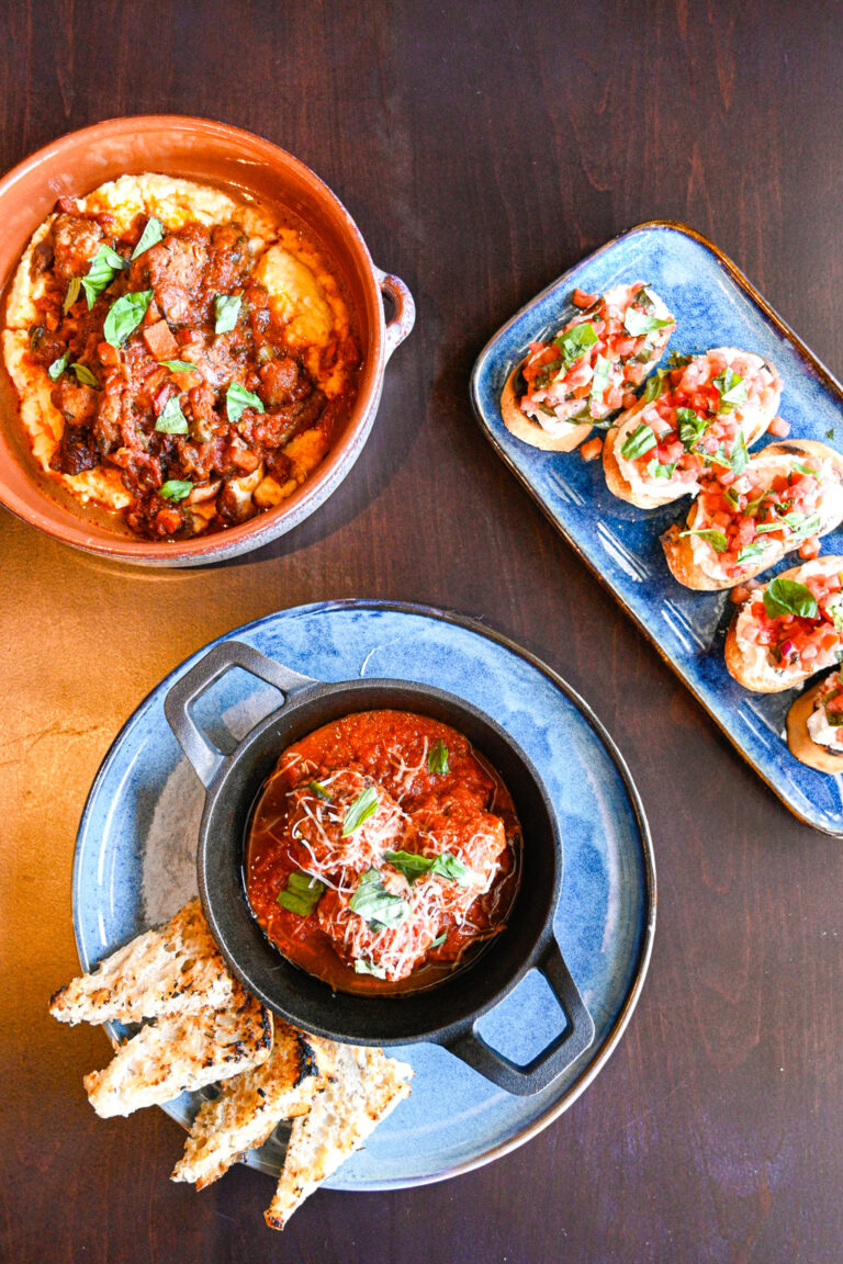 An overhead shot of three dishes on a wooden table. The top dish is a bowl of tomato-based stew with meat and herbs. The bottom plate has a smaller bowl of red sauce with cheese, garnished with basil, and sliced bread on the side. The right dish is a platter of bruschetta.