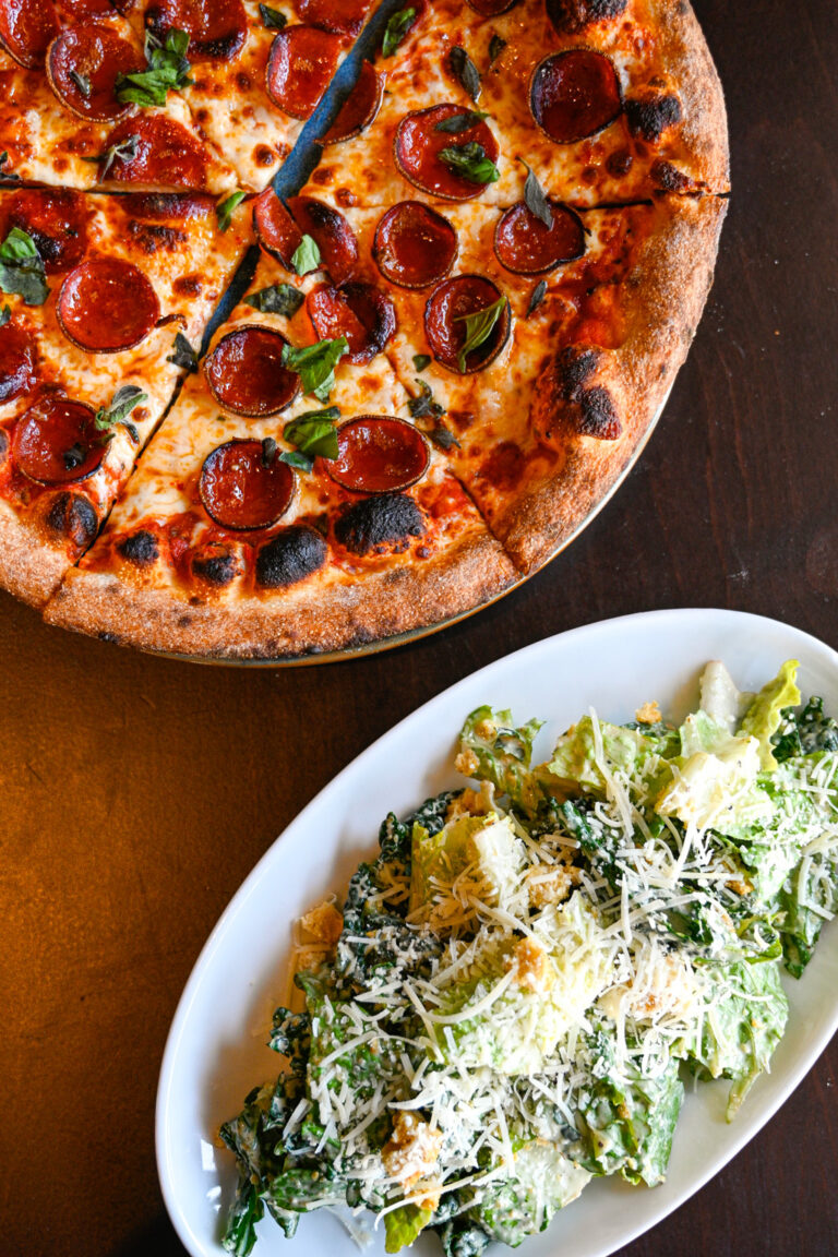 A pepperoni pizza with fresh basil sits on a dark wooden table next to a white oval plate filled with Caesar salad topped with grated cheese.