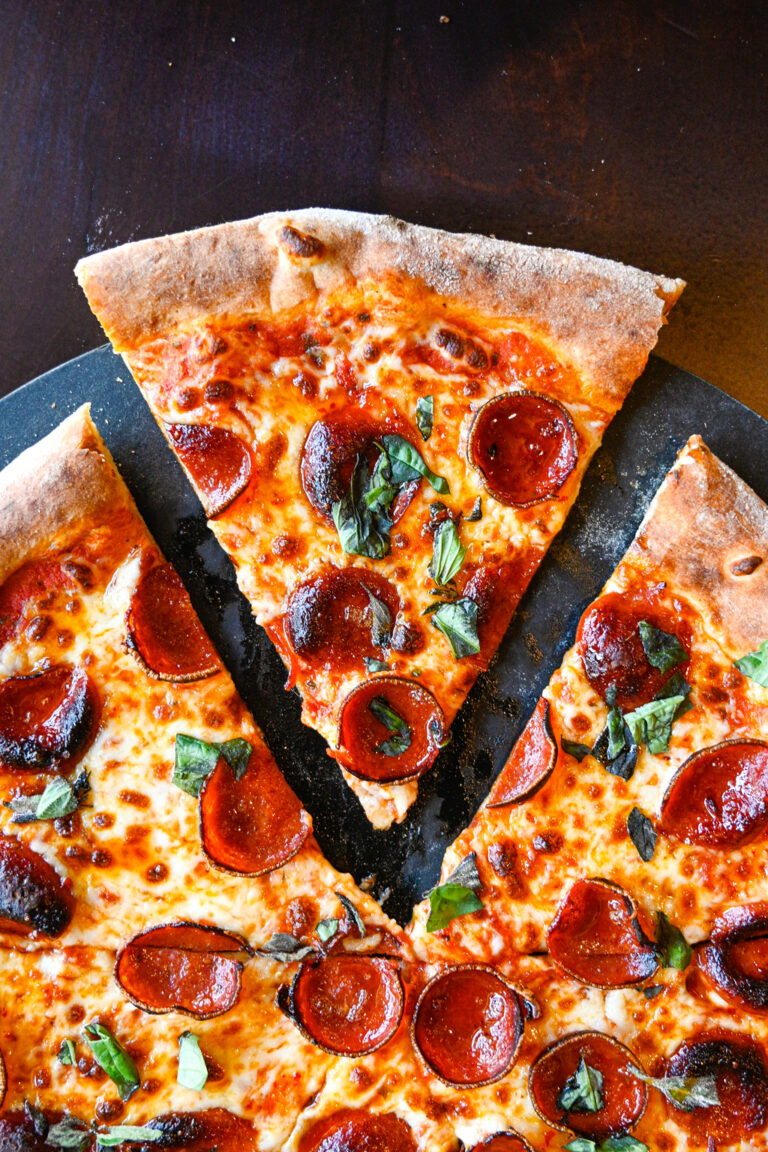 A close-up of a pepperoni pizza with one slice slightly pulled away from the rest. The pizza is topped with melted cheese, pepperoni slices, and fresh basil leaves. The crust appears crispy and golden brown.