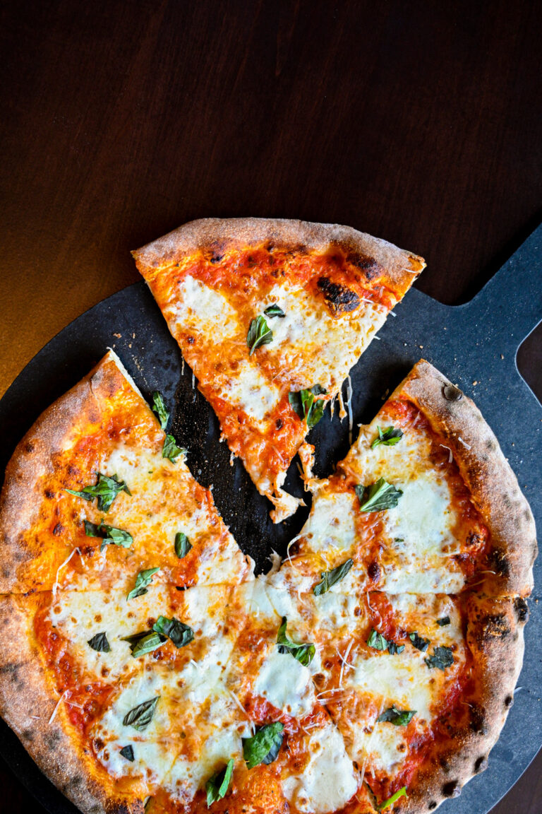 A freshly baked margherita pizza is displayed on a dark surface. One slice is pulled away, revealing melted cheese and fresh basil leaves on a tomato sauce base. The crust is golden brown and slightly charred.