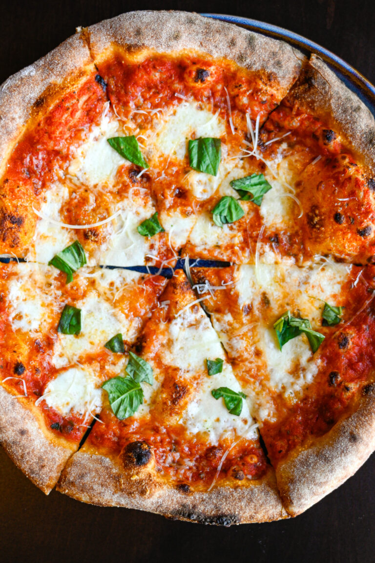 A delicious, freshly-baked pizza with a golden-brown crust, topped with melted mozzarella cheese, dollops of tomato sauce, and fresh basil leaves scattered across. The pizza is cut into six slices and placed on a dark surface.