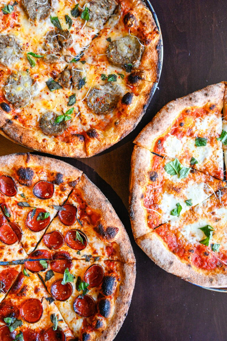 Three pizzas on a dark wooden table. The top left pizza has meatball slices and basil, the bottom left pizza is topped with pepperoni and basil, and the right pizza has a margherita style with mozzarella, basil, and tomato sauce.