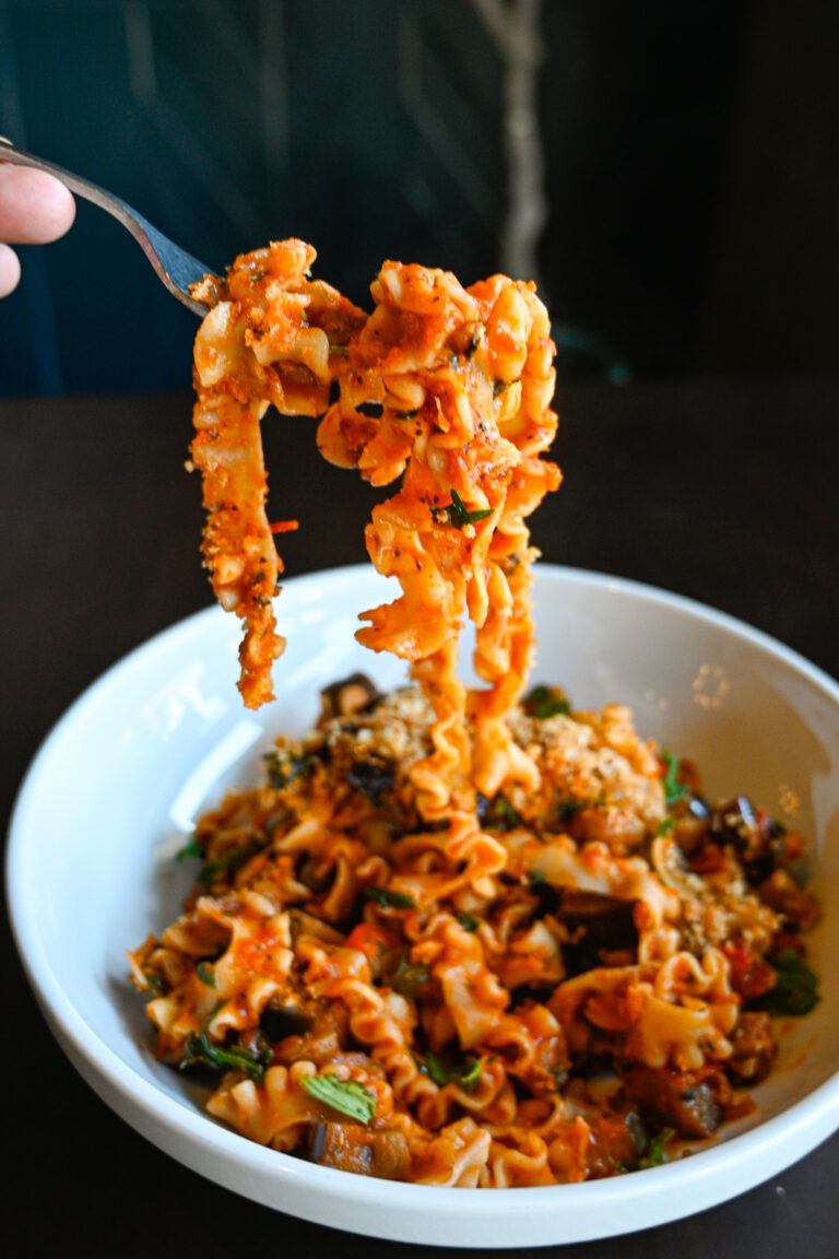 A fork lifts a twirl of wide, curly pasta coated in a rich tomato sauce from a white bowl. The bowl is filled with the same pasta mixed with vegetables and herbs, creating a colorful and appetizing dish ready to be enjoyed.