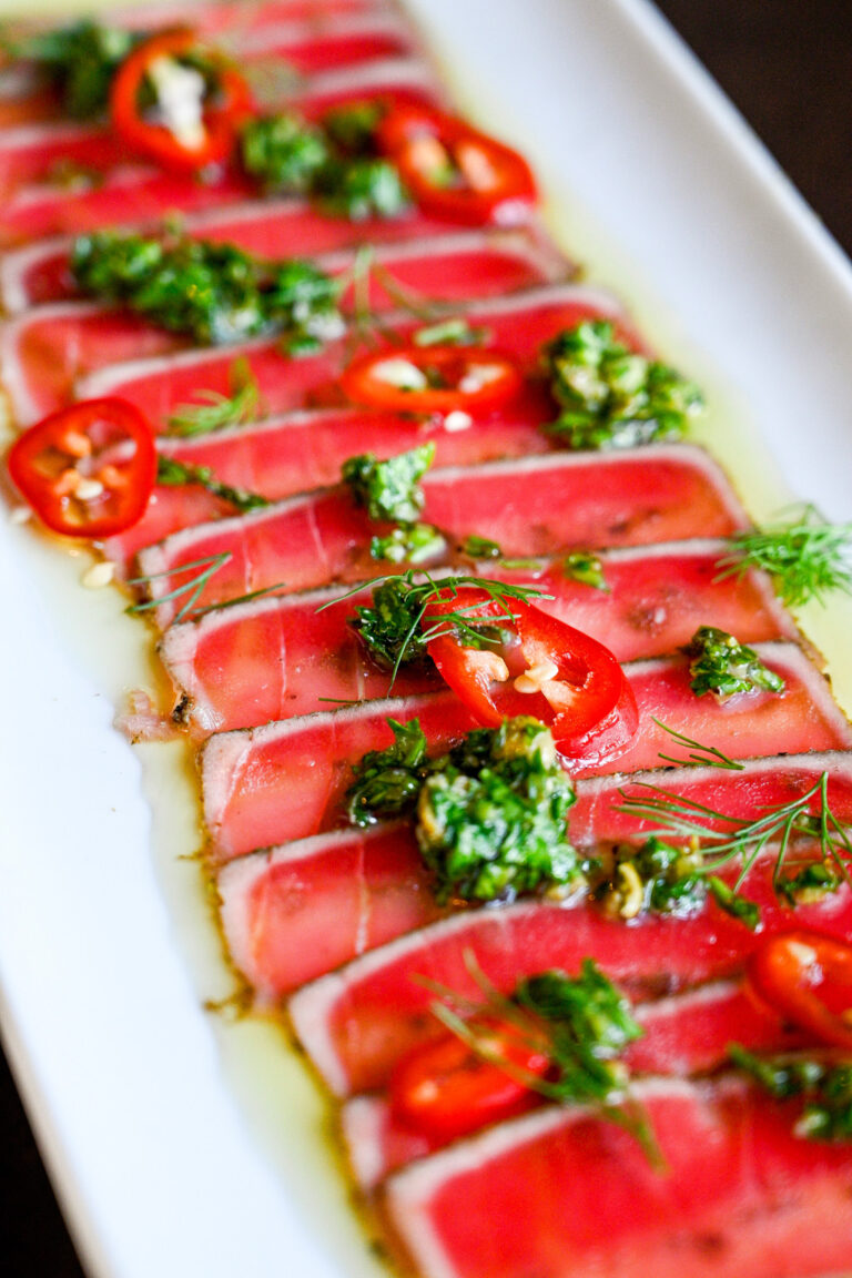 A white rectangular plate featuring thinly sliced seared tuna topped with fresh herbs, chopped garlic, and sliced red chili peppers, drizzled with a green sauce. The tuna is arranged in a neat, overlapping pattern.