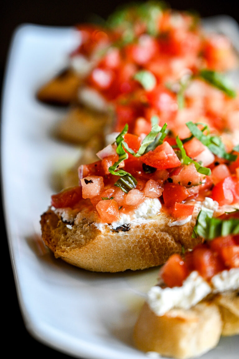 A close-up of bruschetta on a white plate. The toasted bread slices are topped with chopped tomatoes, fresh basil, and a spread of ricotta cheese. The vibrant colors and textures make the dish look fresh and appetizing.